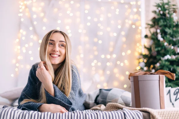 Το κορίτσι ξαπλώνει στο κρεβάτι και χαμογελάει. Χριστουγεννιάτικο δέντρο και γιρλάντες κατά την t — Φωτογραφία Αρχείου