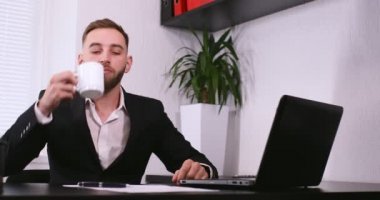 İş adamı büro masasında oturuyor, kahve içiyor ve dizüstü bilgisayar başında çalışıyor.