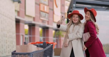 Neşeli genç kadınlar alışveriş merkezinin yakınındaki kağıt alışveriş torbalarıyla selfie çekiyorlar. En iyi arkadaşlar şık kıyafetler ve şapkalar giyerler..