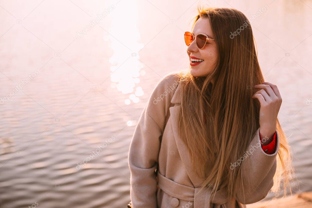 Portrait of a young smiling girl in coat on a background lake at sunset