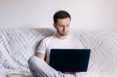Genç ve çekici bir adam evdeki kanepede oturuyor internette dizüstü bilgisayarla çalışıyor, internet kullanıyor, boş zaman geçiriyor, rahat bir hayat sürüyor.