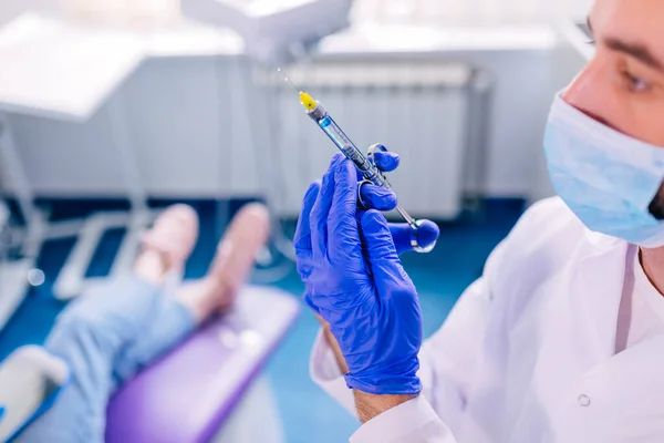 the dentist holds an anesthesia syringe against the background of his patient sitting in a dental chair.