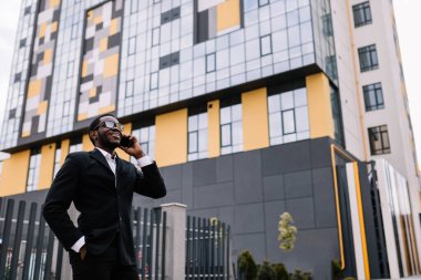 Gülümseyen genç Afrikalı Amerikalı iş görüşmesi yapıyor, sokakta telefonla konuşuyor..