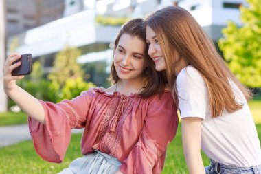 İki liseli kız, yazın bir parkta, akıllı telefondan selfie çekiyorlar, günlük kıyafetler giyiyorlar, okuldan ve üniversiteden bir süre uzaklaşıyorlar.,