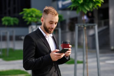Tek kullanımlık kahve bardağıyla resmi takım elbiseli genç adam akıllı telefon ekranına bakıyor ve gülümsüyor.