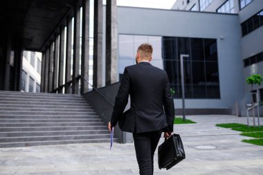 Resmi takım elbiseli, evrak çantalı genç adam ofis binasının önünden geçiyor.