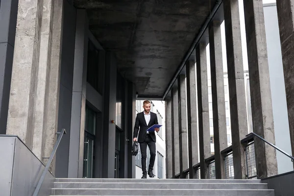 young man in formal suit walking by office building with briefcase and clipboard