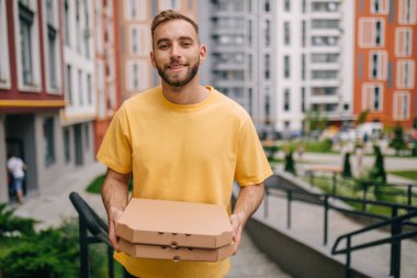Sarı tişörtlü bir teslimatçının elinde iki paket pizza kutusuyla kameraya baktığı fotoğraf..