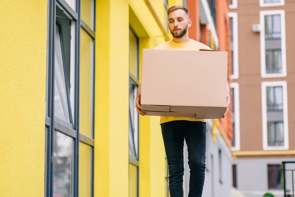 The delivery man carries a box. A man delivers goods to consumers. A large brown cardboard box in the hands of a worker.