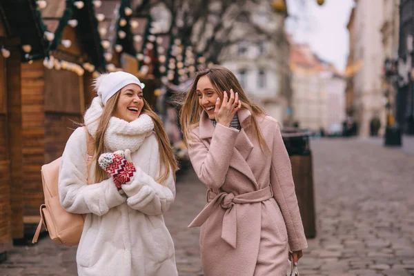 ニットで買い物をしている2人の友人は 冬の日にガーランドで飾られた市場の広場を歩いています 冬のお祝い気分をお楽しみください — ストック写真