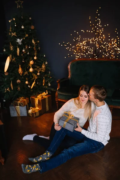 Handsome man in a sweater holds a large box, gives it to the girl who sits in a cozy living room near a shiny garland on a pine tree