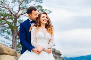 Damadın yakın çekimi, gelini arkadan sarar muhteşem bir manzarayla kayalıklarda.