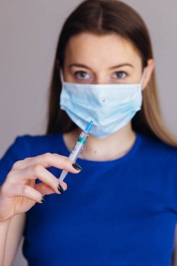 Maskeli bir kadın doktor elinde şırıngayla arka planda bulanık bir kadının şırıngasını tutuyor.