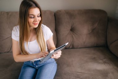 Çekici genç bir kadın dijital tablet kullanıyor ve evdeki kanepede otururken gülümsüyor.