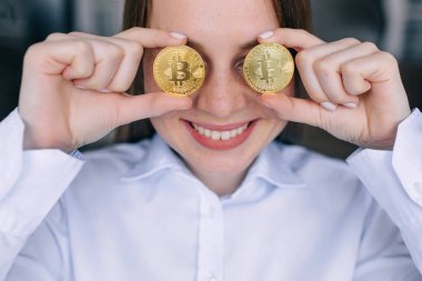 Genç, mutlu, gülümseyen bir kadın elinde gözlük yerine iki adet bitcoin tutuyor.
