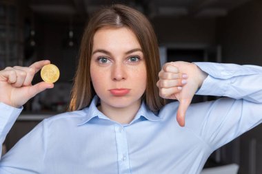 Elinde kızgın yüzüyle Bitcoin tutan genç bir iş kadını. Negatif işaretler baş parmakları aşağıda hoşnutsuzluk gösteriyor.
