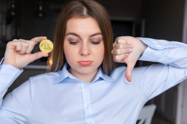 Elinde kızgın yüzüyle Bitcoin tutan genç bir iş kadını. Negatif işaretler baş parmakları aşağıda hoşnutsuzluk gösteriyor.