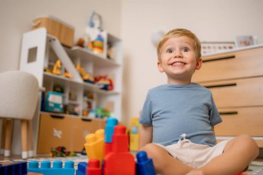 Cheerful young boy sitting on the floor surrounded by colorful toys in a bright playroom setting.