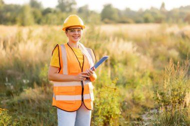 A cheerful professional woman engineer inspecting a field in safety gear holding documents , surveying an outdoor field setting.