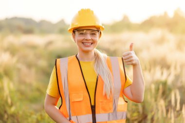 A confident construction worker wearing a yellow hard hat and safety vest gives a thumb-up in an outdoor setting during a sunny day, enthusiasm, positivity, and adherence to safety measures.