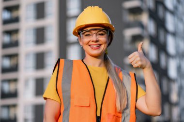 Modern construction worker in safety gear smiling and showing positivity giving thumb up near city skyscrapers.