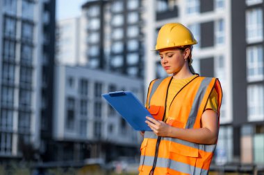 A woman on a construction site dressed in a reflective vest and helmet checks documents carefully, representing workplace safety, professionalism, and progress in an urban development context.
