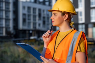 A focused and professional female construction worker wearing safety gear evaluates the project site. Holding a clipboard, reflecting on the progress against modern city building