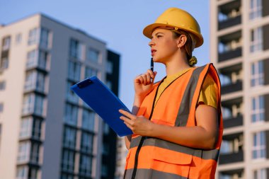 A female construction worker in safety gear holds a clipboard, reviewing plans near contemporary apartments.