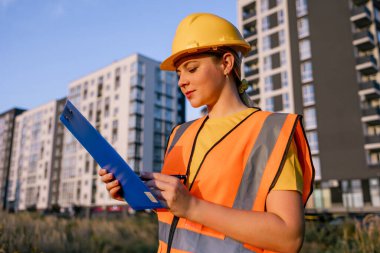 A construction worker in safety gear evaluates documents on-site with urban skyline in the background, emphasizing planning and readiness in a modern construction environment.