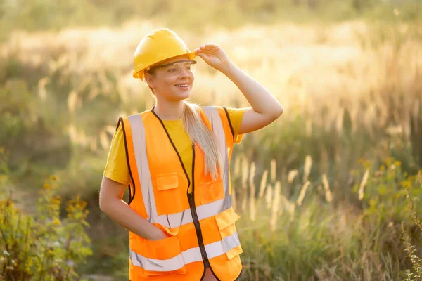 A cheerful female worker in safety gear, a helmet and vest, standing outdoors in a grassy field under sunlight, demonstrating positivity and professionalism often attributed to the confident workforce.