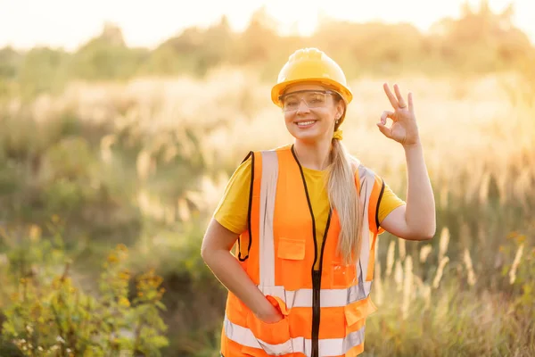 A happy female construction worker wearing safety gear in a summer field, gesturing an okay sign, conveying positivity and professionalism in an outdoor work environment.