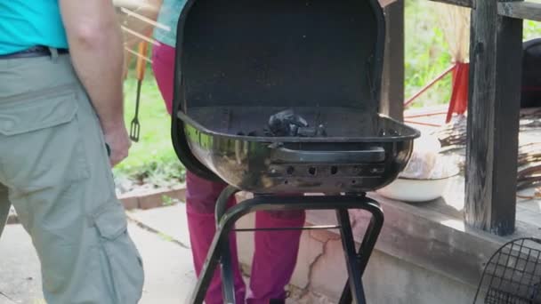 Um homem gosta das brasas no churrasco, e uma mulher ajusta as brasas com pinças. — Vídeo de Stock