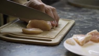 Bir adam büyük bir bıçakla bir tavuk filetosunu doğruyor.