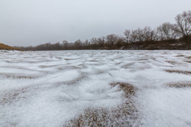 Vahşi bir nehir kıyısındaki soyut kum tepecikleri, taze kar tozuyla kaplı.