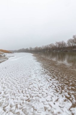 Vahşi bir nehir kıyısındaki soyut kum tepecikleri, taze kar tozuyla kaplı.