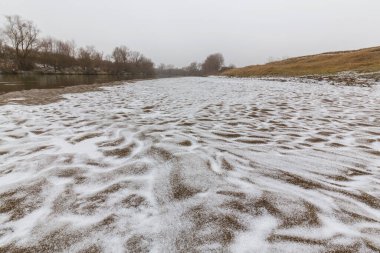 Vahşi bir nehir kıyısındaki soyut kum tepecikleri, taze kar tozuyla kaplı.