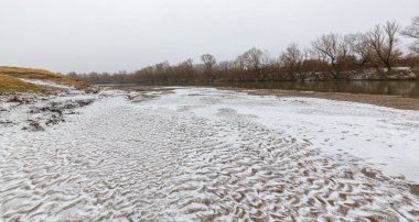 Vahşi bir nehir kıyısındaki soyut kum tepecikleri, taze kar tozuyla kaplı.