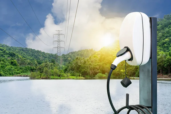 Ev station plug and power cable supply for electric vehicle charging or hybrid car on natural blue sky sunshine and high voltage electric pole at river and green mountain background