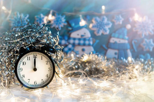 New Year alarm clock with silver tinsel on top and blue background with snowmen behind. Midnight.