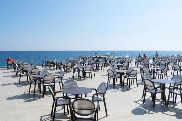 Yaz zamanı. Deniz manzaralı açık hava kafe. Deniz kenarında siyah kenarlı beyaz masalar ve sandalyeler. Yatay yönelim.