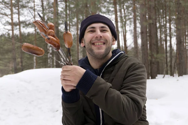 Young smiling man holding a sausage on a spit in the woods