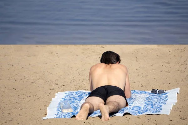 Glad tanned man with closed eyes lies with his hands under the head on the sand beach outdoors. Sun shines at his body. He wears white wireless earphones and a dark watch. — Stock Photo, Image