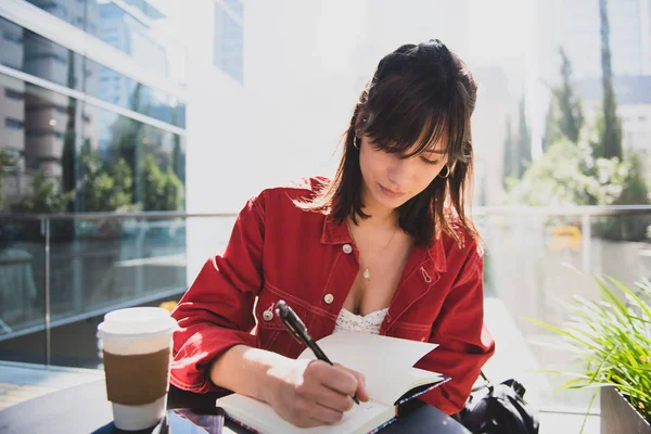 Young happy woman at a coffee shop outdoors writing on a book