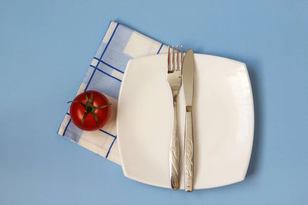 Fourchette, couteau et assiette blanche avec tomate sur fond bleu. Assiette propre et couverts sur fond clair. Vue du dessus. — Photo