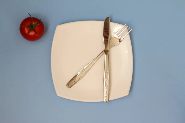 Fourchette, couteau et assiette blanche avec tomate sur fond bleu. Assiette propre et couverts sur fond clair. Vue du dessus. — Photo