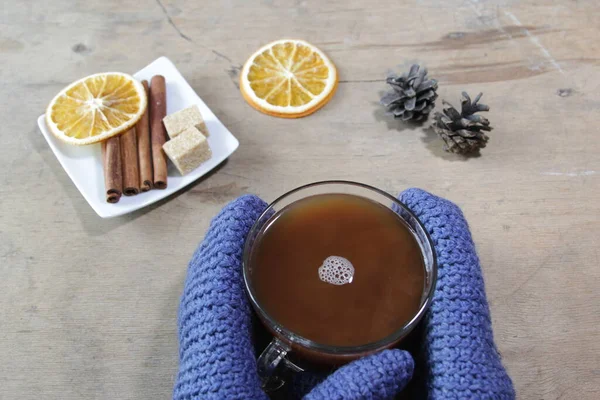 Осенняя или зимняя уютная композиция. Чашка какао, коричные палочки, сахар, апельсин и сосновые шишки на деревянном фоне. Руки в синих рукавицах с чашкой какао. — стоковое фото