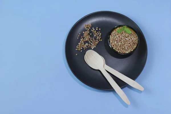 Buckwheat grains on black plate and buckwheat in a bowl, blue background. Gluten free grain for healthy diet. Selective focus. Top view. Copy space. — Stock Photo, Image
