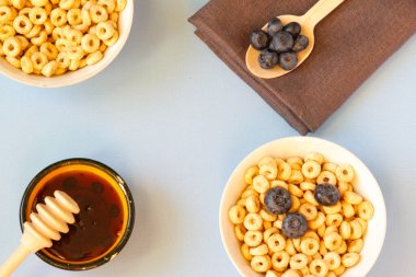 Bal, mısır gevreğini süt ve yaban mersiniyle masaların mavi arka planında beyaz bir kaseye koyar. Sağlıklı kahvaltı. Uzayı kopyala Yatay oryantasyon. Üst Manzara. Düz yatıyordu