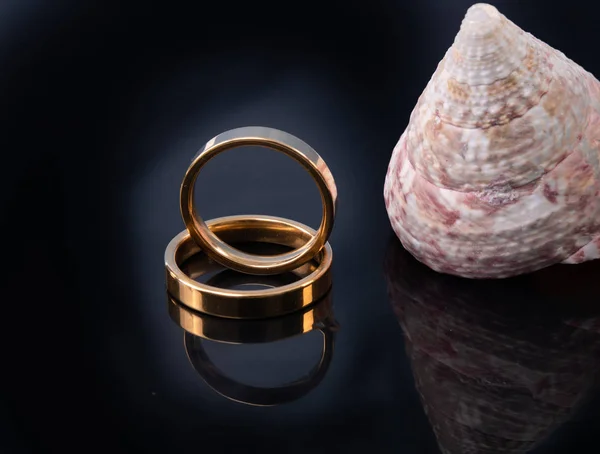 Ragyogó arany jegygyűrű egy sötét felületre kíséri a tengeri kagyló. Jegygyűrűk és sablon szöveg — Stock Fotó