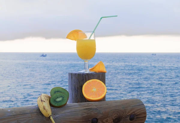 Green kiwi slices, ripe orange slices, banana and a delicious orange juice - Boats in the blue sea under cloudy sky background
